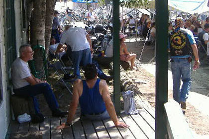 Men sit on porch and crowds of people sit at eating tables in the distance.