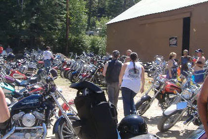Parking lot filled with motorcycles and a few people.