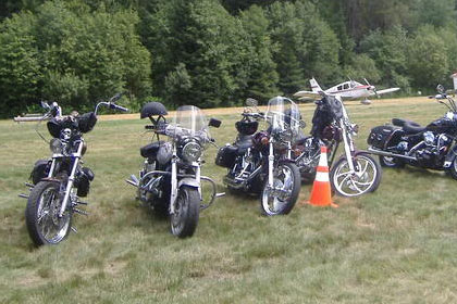 Line of motorcycles parked in grass with small airplanes parked in background.
