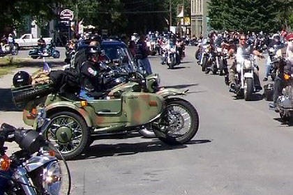 Motorcycles line both sides of the street while more cycles drive towards the camera.