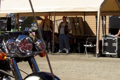 Motorcycle parked in foreground. Men stand in and around tent in background.