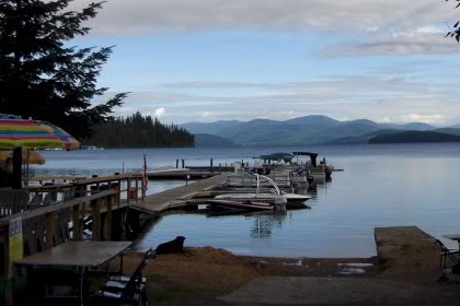 Tables set up by lake shore. Dock with boats, trees, and mountains in the distance.