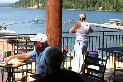 Several groups of people sit around tables. Lake and dock filled with boats in the background.