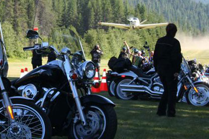 Line of motorcycles parked in grass with small airplane coming in for landing in background.