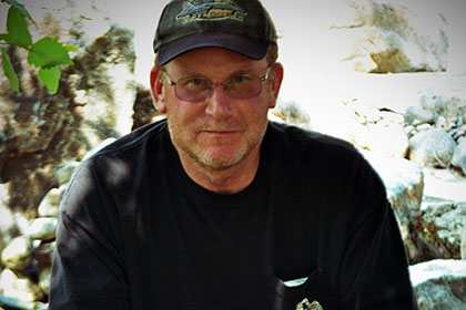 Man with hat and glasses sitting on a large rock.