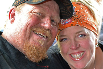 Bearded man with hat and a woman with an orange bandana, smiling for camera.