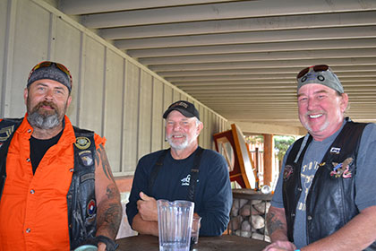 Three smiling guys sitting at a table outside.